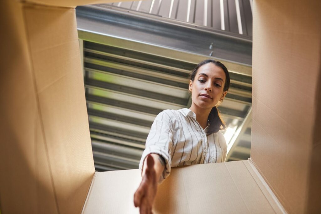 A woman in a white and gray striped shirt reaches into a cardboard box while the camera points up at her from the bottom of the box. She is in an industrial setting.
