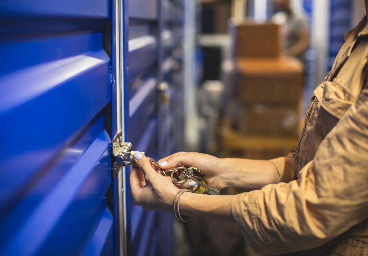 A woman in a tan jacket uses a ring of keys to unlock a blue storage unit door and a stack of boxes sits behind her.