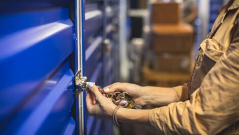 A woman in a tan jacket uses a ring of keys to unlock a blue storage unit door and a stack of boxes sits behind her.