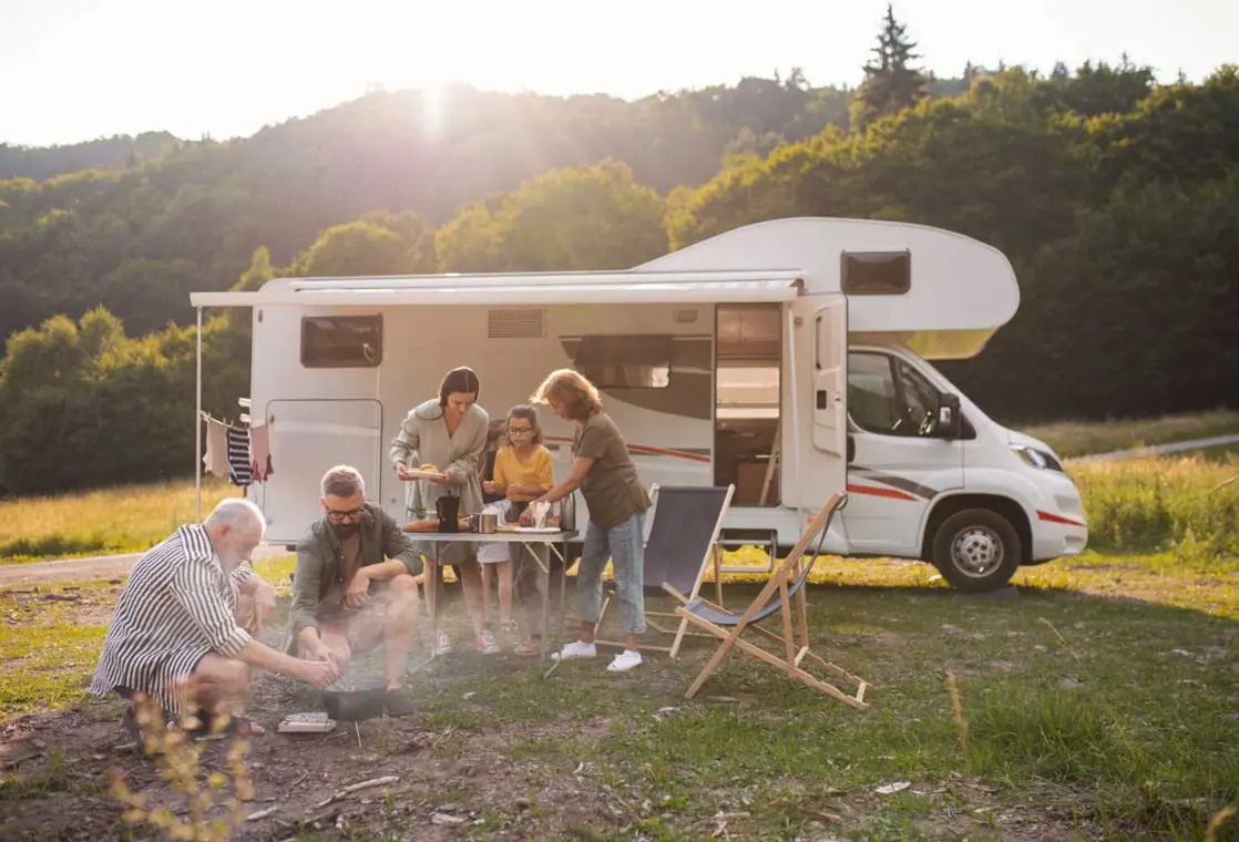 A family gathers outside of a camper at a folding table by a fire