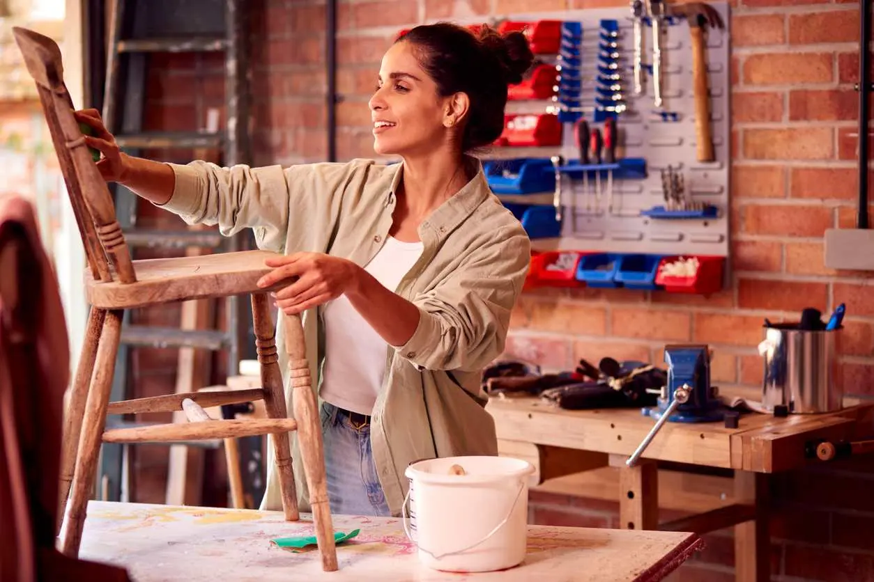 A woman sands a wooden chair that is sitting on a workbench in a garage