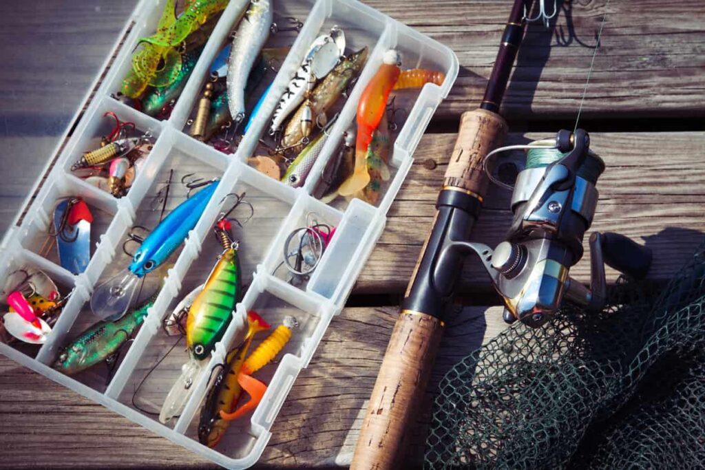 Fishing lures inside a tackle box next to a fishing rod and net on a wooden pier.