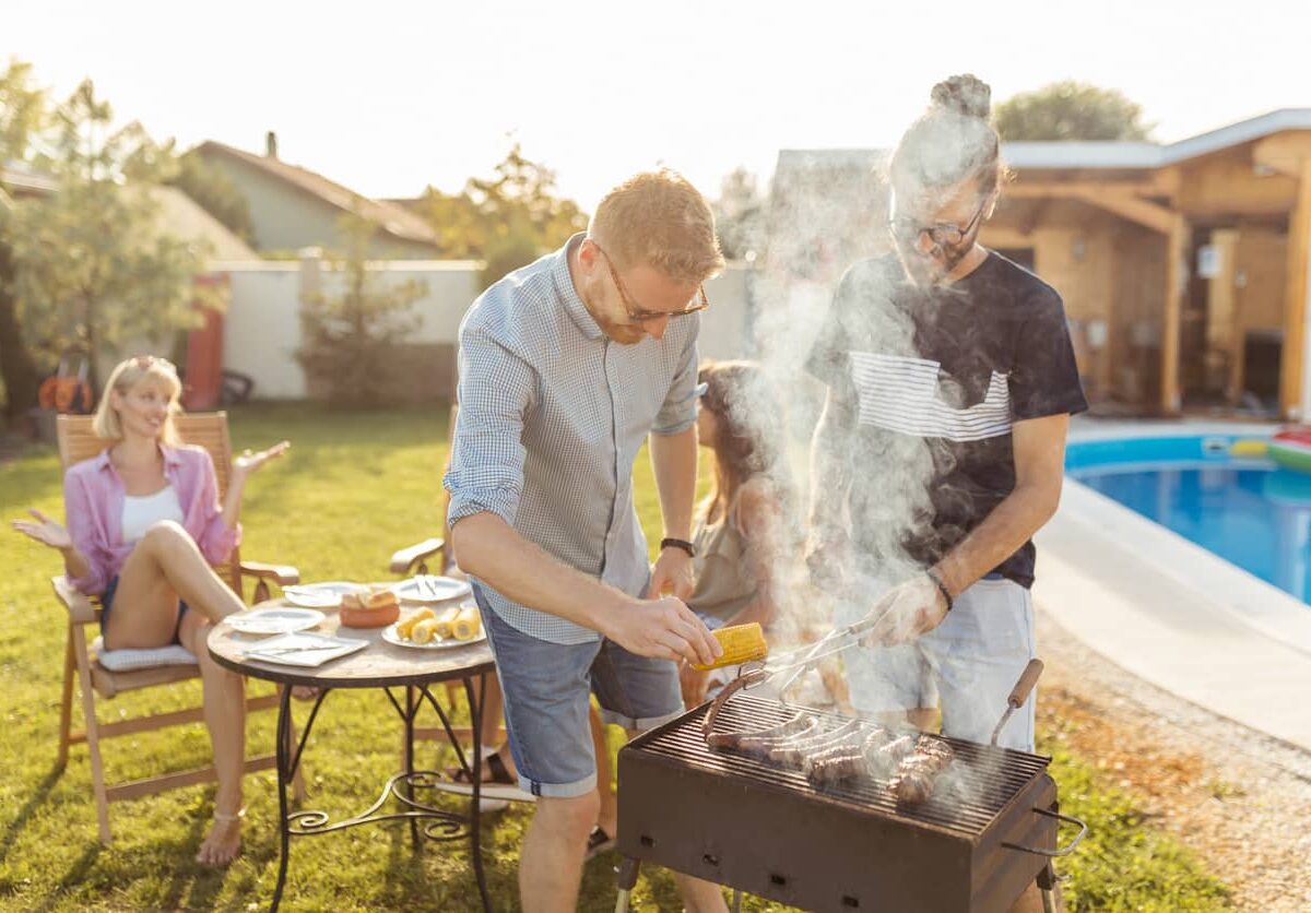 Two men cooking on a small bbq grill while two women sit around a table in the background.