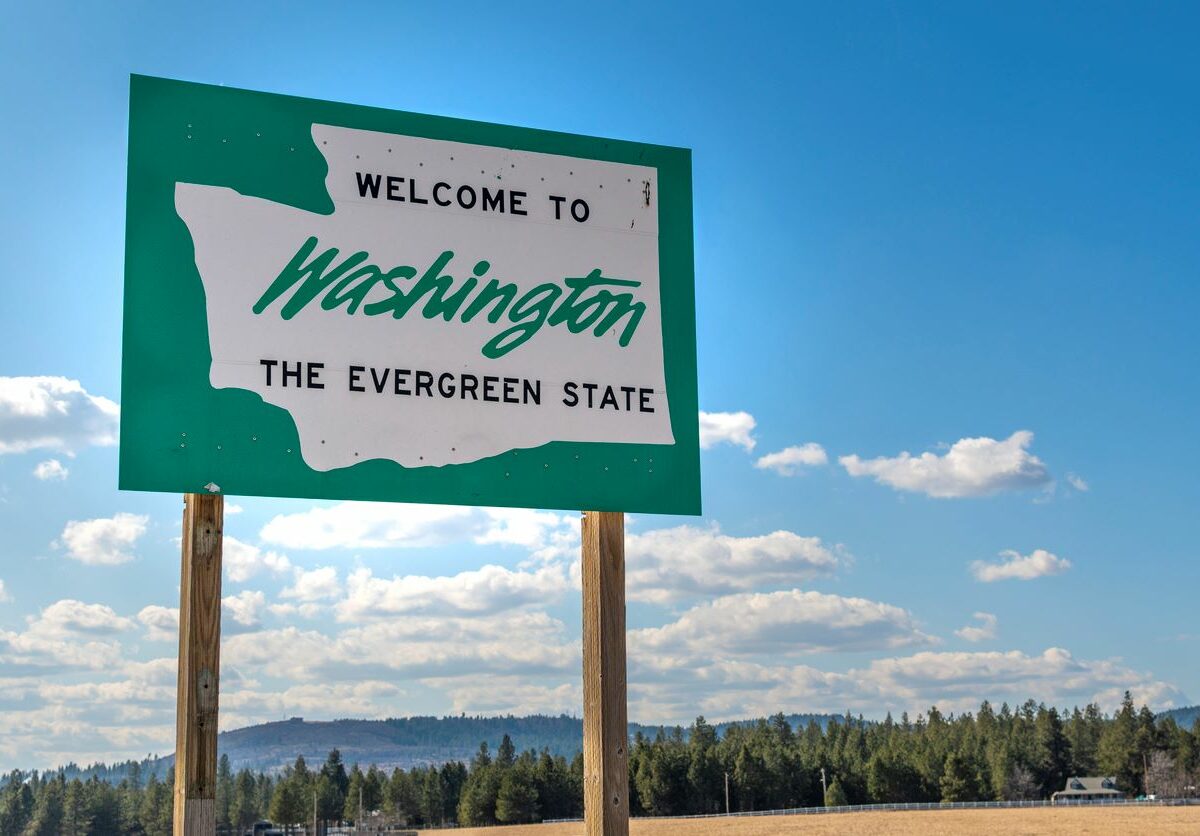 A green sign features the shape of Washington with text reading, “Welcome to Washington, the Evergreen State,” with a backdrop of a partly cloudy sky and evergreen trees.