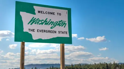 A green sign features the shape of Washington with text reading, “Welcome to Washington, the Evergreen State,” with a backdrop of a partly cloudy sky and evergreen trees.