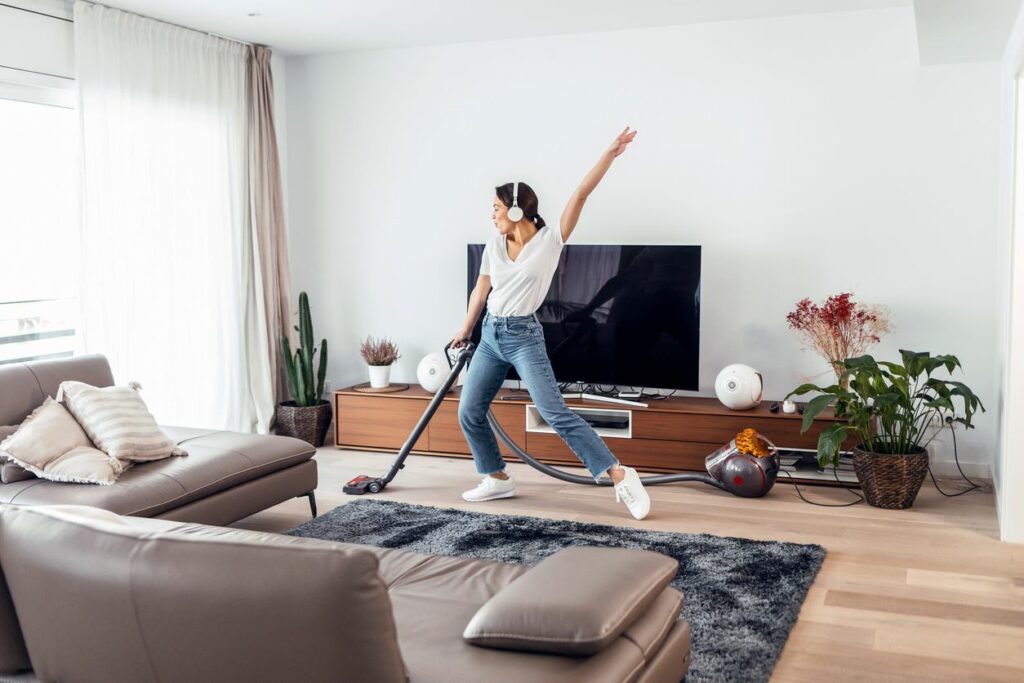 A young, happy woman listening and dancing to music while cleaning the living room floor with a vacuum cleaner.