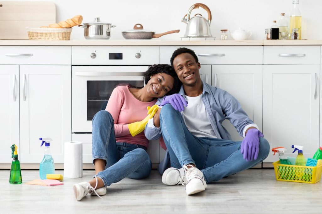  A couple in rubber gloves rests after cleaning the apartment, sitting on the floor in front of the stove in the kitchen among housekeeping supplies.