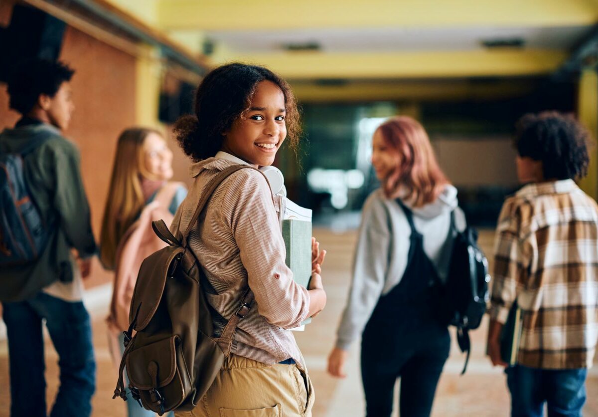 A young girl with a backpack happily walks down a school hallway with friends.