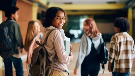 A young girl with a backpack happily walks down a school hallway with friends.