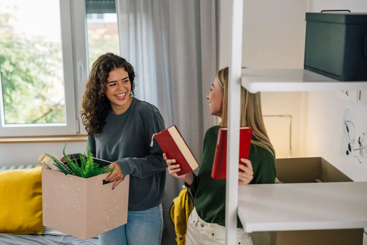 Two female college students move into a dorm room holding books and a box. 