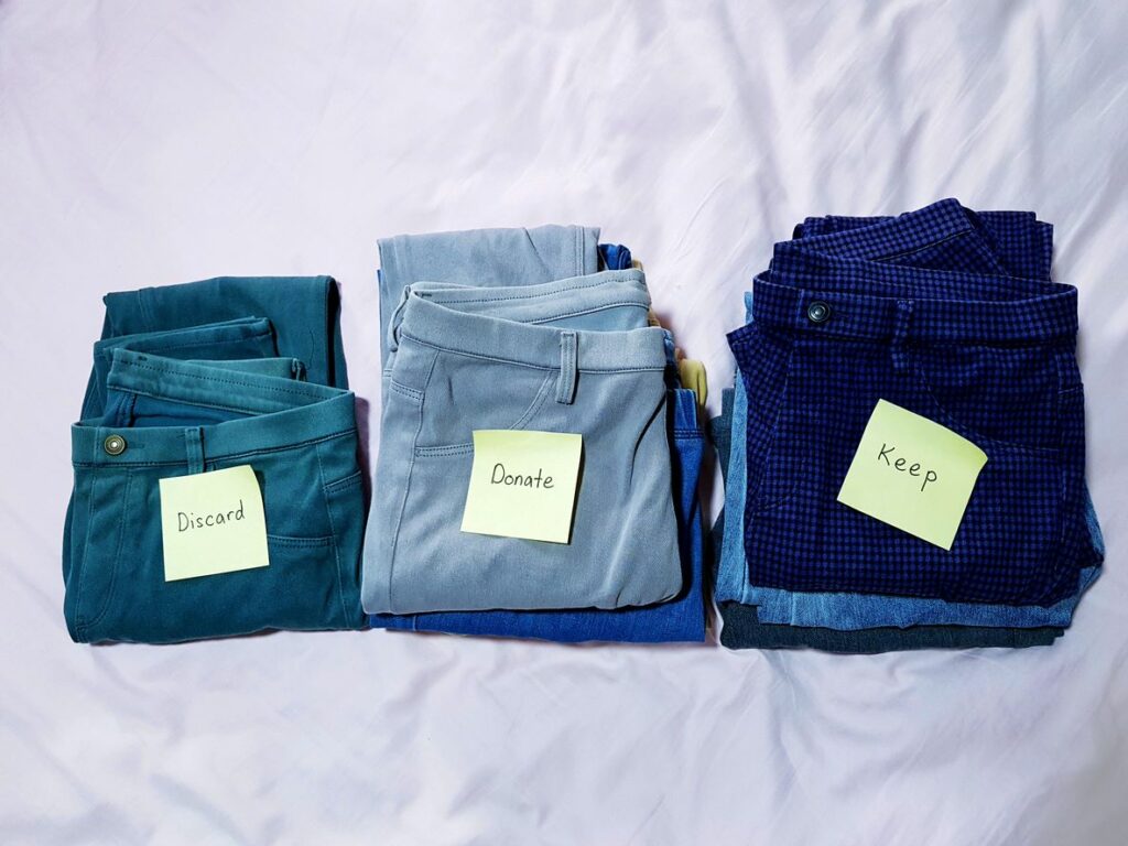 Three stacks of folded pants sit on a bed, with sticky notes reading “discard,” “donate,” and “keep.” 