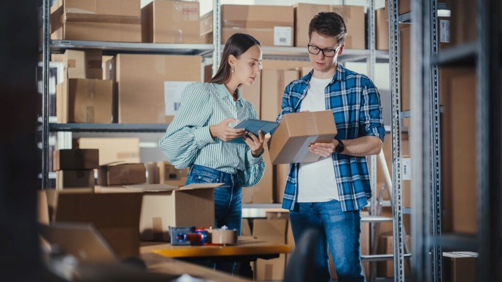 Two young warehouse workers examine a package with shelves of boxes behind them.