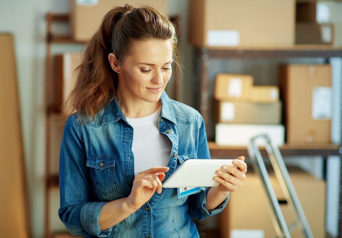 A young woman with a ponytail looks at a tablet with boxes of supplies on a shelf behind her