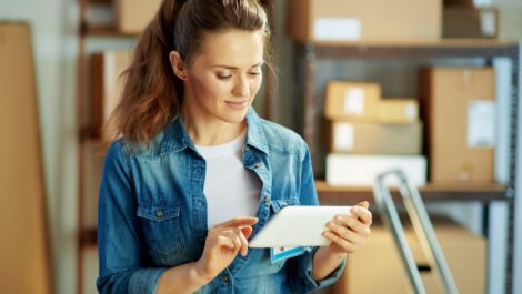 A young woman with a ponytail looks at a tablet with boxes of supplies on a shelf behind her