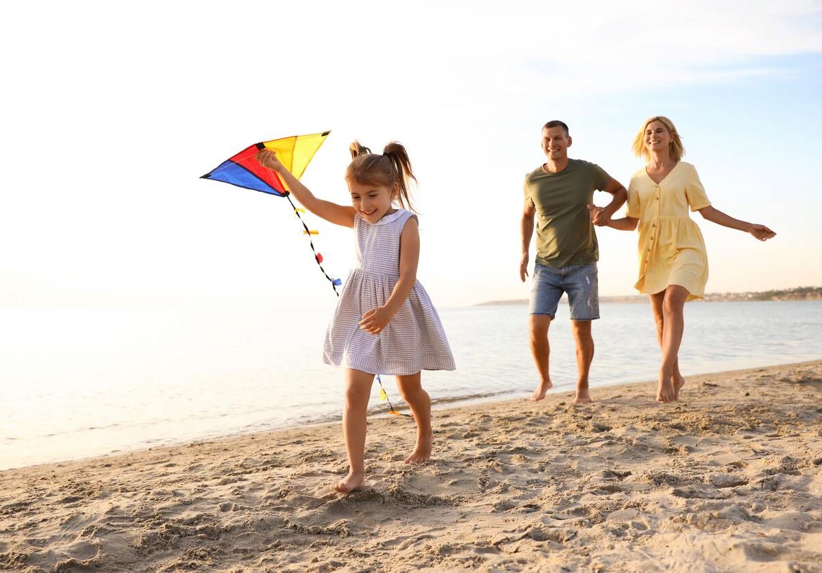 A young daughter holds a kite on the beach as her smiling parents follow behind.