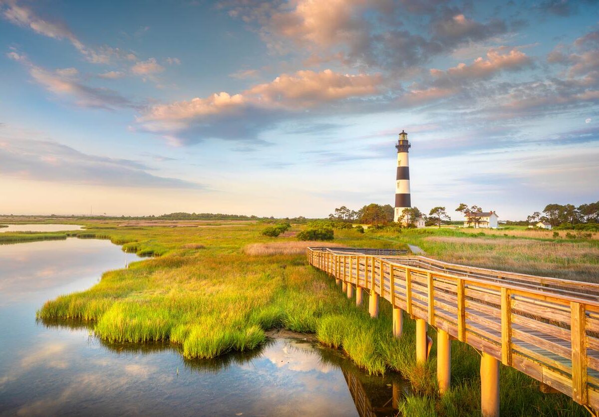 A wooden pier off Cape Hatteras National Seashore with Bodie Island Lighthouse behind at sunrise.