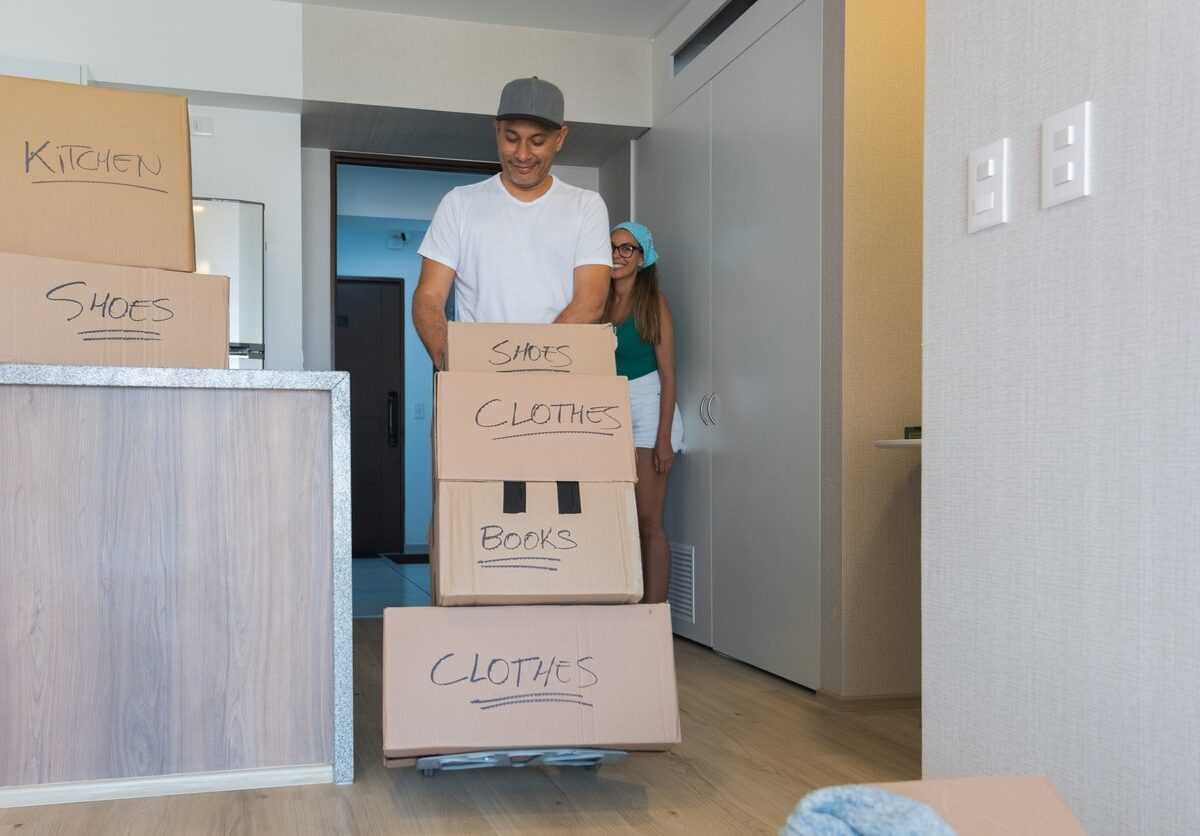 A man pushes a dolly with labeled boxes, and a woman holds the door as they move into their new apartment.