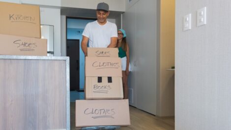 A man pushes a dolly with labeled boxes, and a woman holds the door as they move into their new apartment.