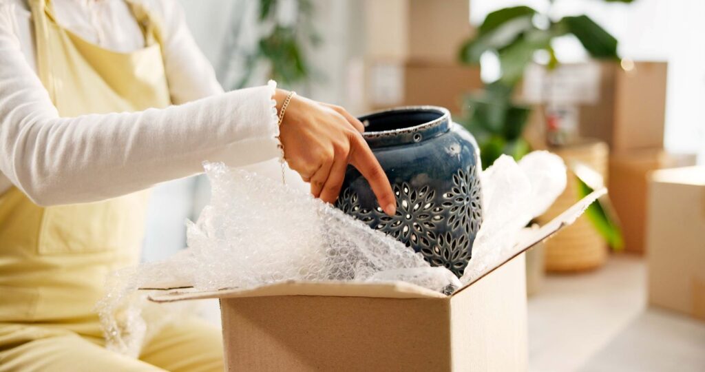 A person in yellow removes a fragile vase from a cardboard box with Bubble Wrap surrounding it. 