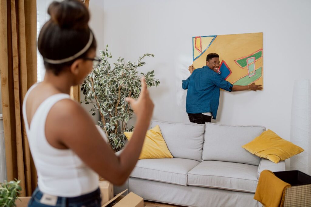 A young woman instructs a smiling young man on where to hang an abstract painting in their new home. 