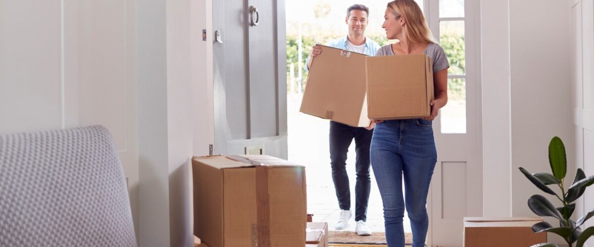 A couple carrying cardboard boxes through the front door of their new home.