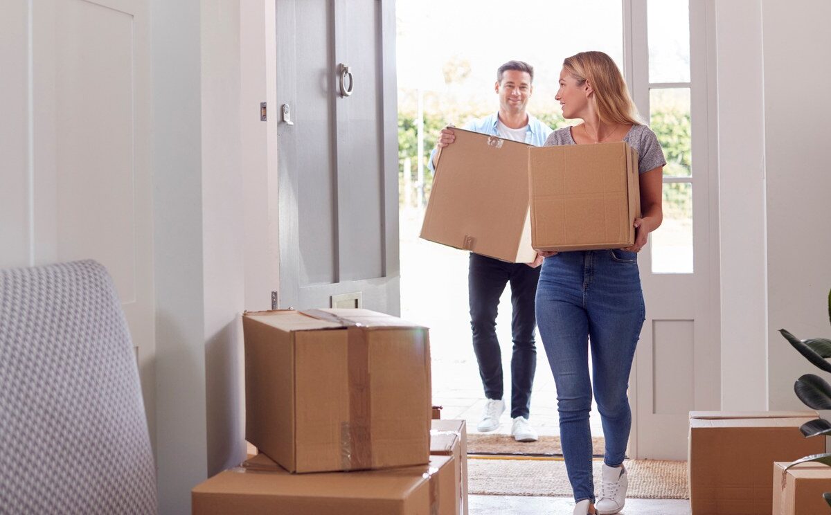 A couple carrying cardboard boxes through the front door of their new home.