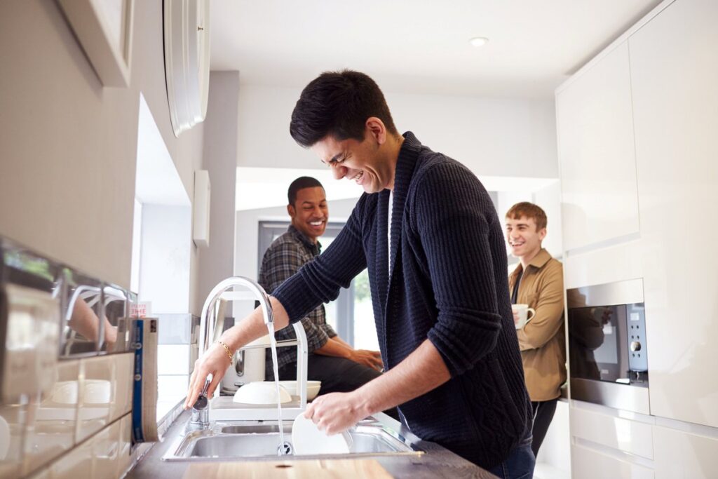 A group of three young roommates laugh as one of them does the dishes. 