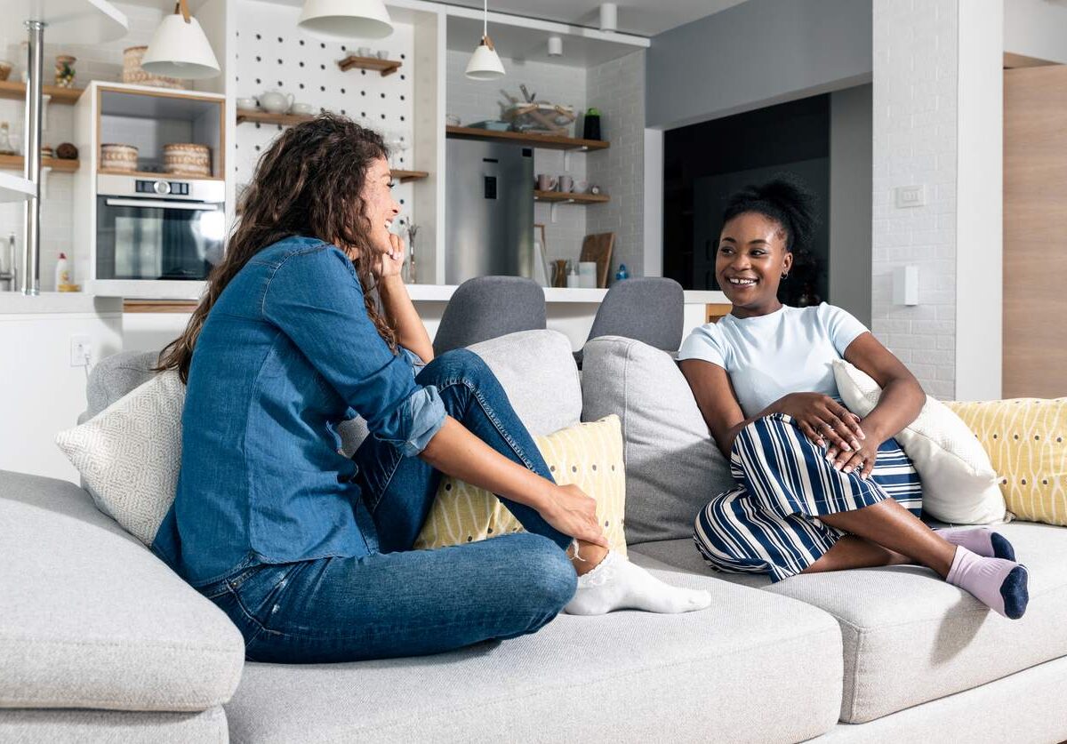 Two smiling young roommates chat on the couch in a living room.