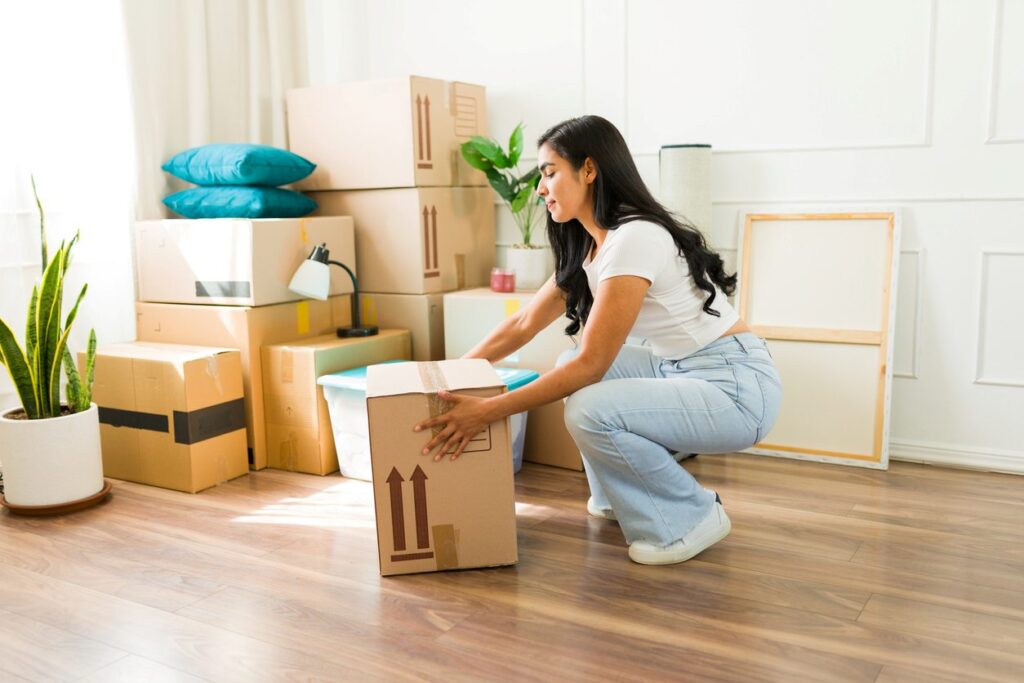 A young adult with long, dark hair sets a box on the wood floor while moving into a new home. 
