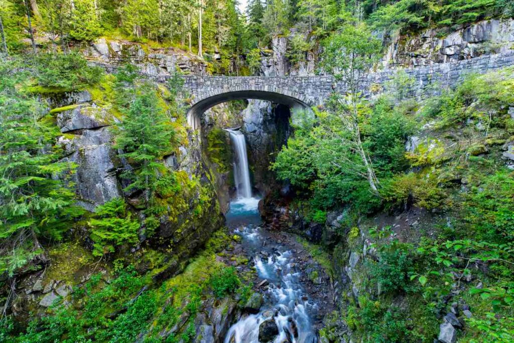 A waterfall framed by a stone bridge in Mount Rainier National Park, surrounded by lush greenery and rocky terrain.