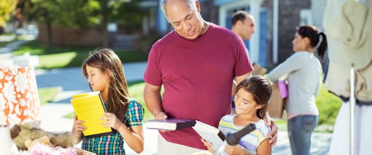 A grandfather and his two granddaughters browse books at a neighborhood garage sale.