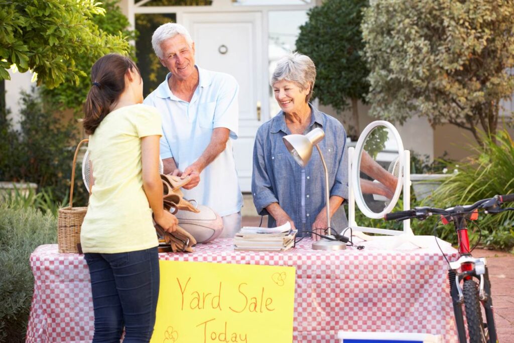 A smiling elderly couple running a yard sale, talking with a young woman at a table. 