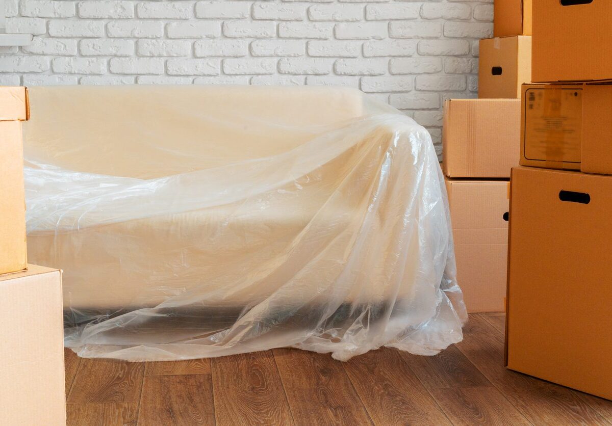 A couch draped in a plastic cover, surrounded by cardboard boxes, sits against a white brick wall.
