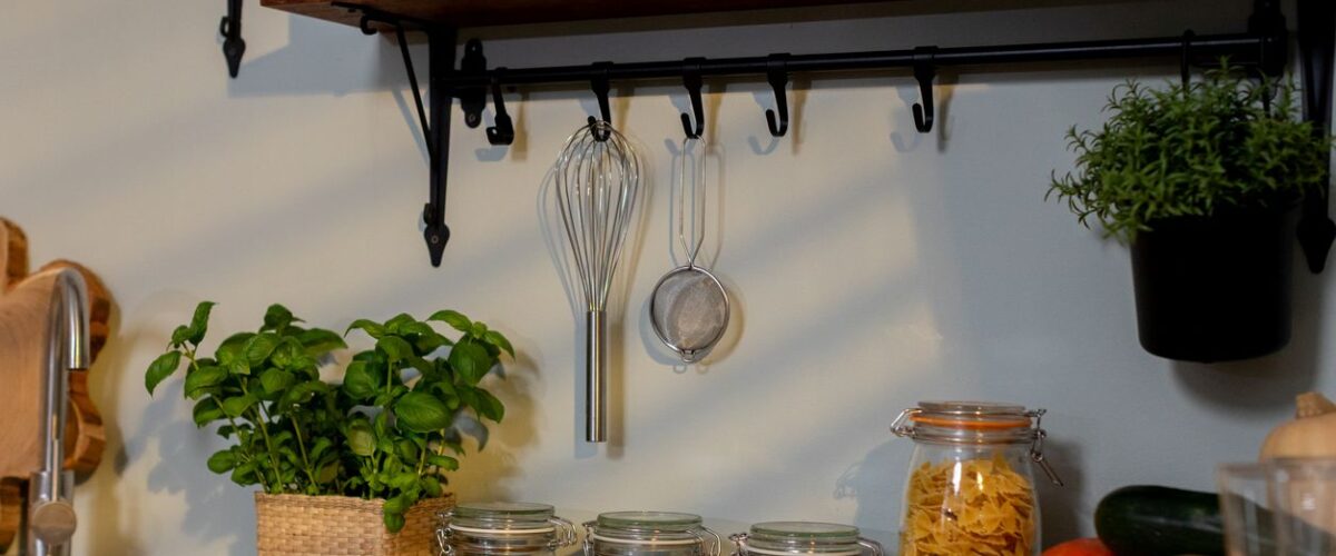 A kitchen filled with jars of food, herb plants, a whisk, a strainer, and rustic decor.