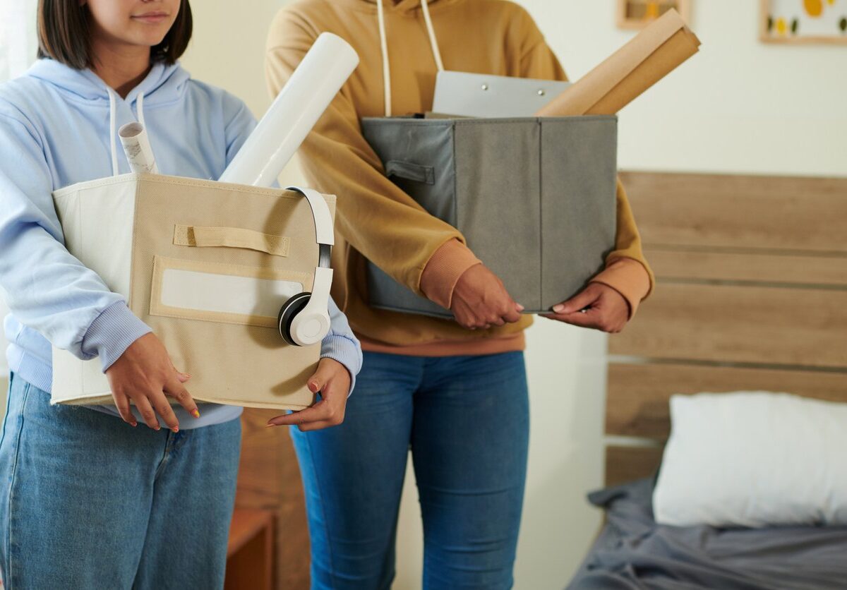 Two people holding cloth bins full of supplies in front of a bed with a wooden headboard and gray sheets.